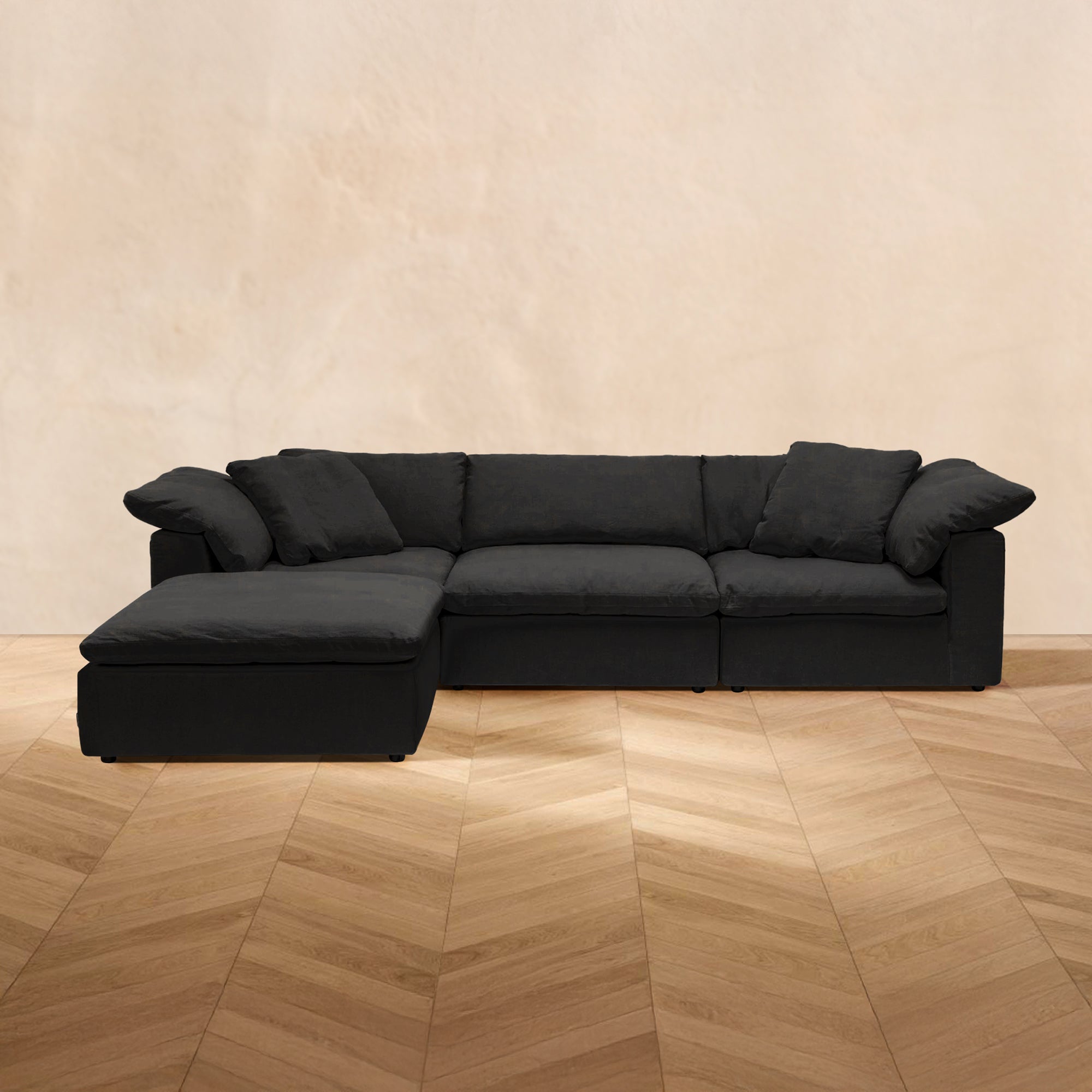 A black HavenlyCloud® 3-Seater L-Sectional Couch with performance fabric cushions and removable covers sits on a wooden herringbone floor against a beige wall.